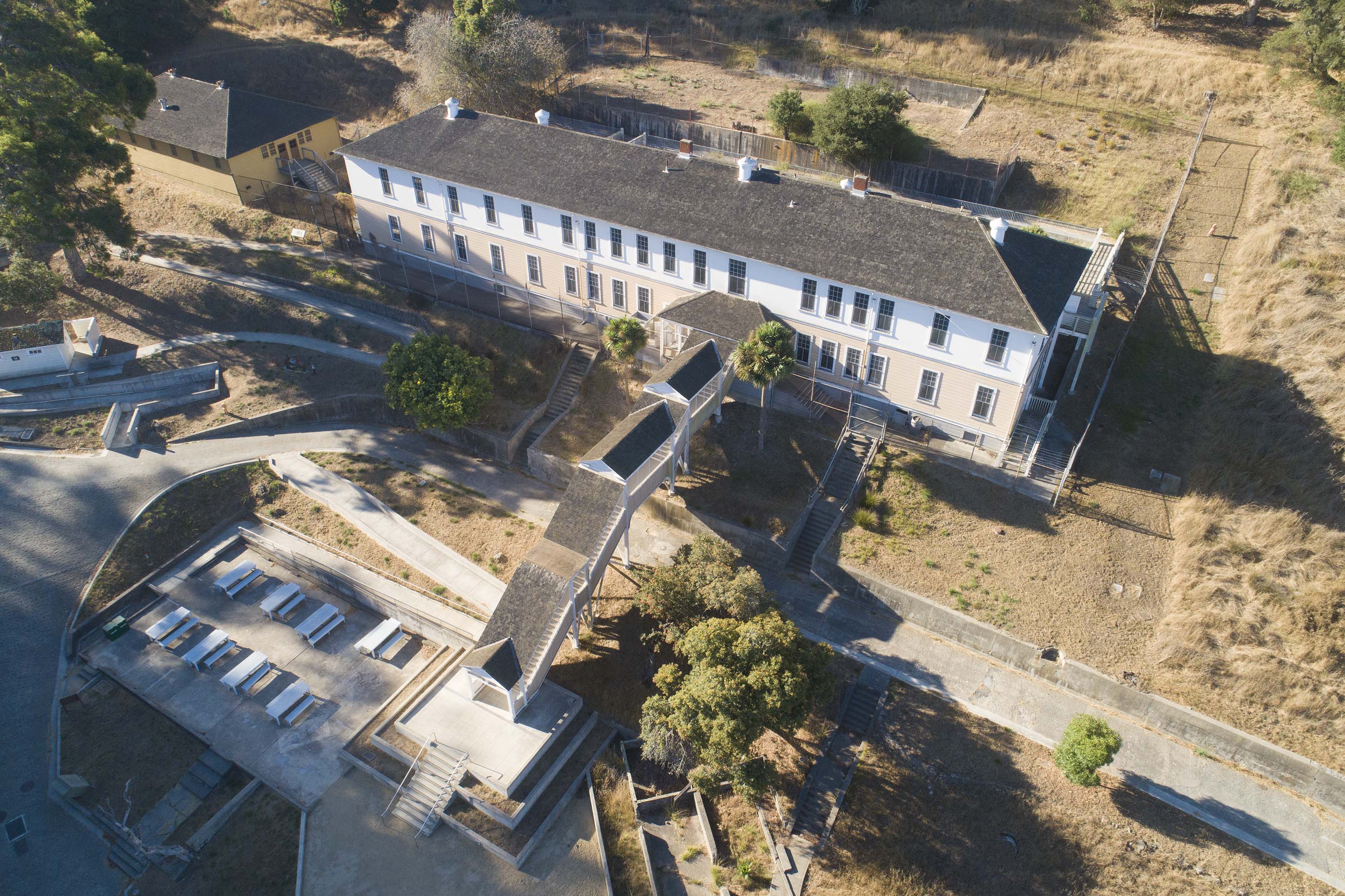 Overhead view of two-story barracks building and concrete foundations built into a sloping hill with eucalyptus trees and other green vegetation surrounding. 