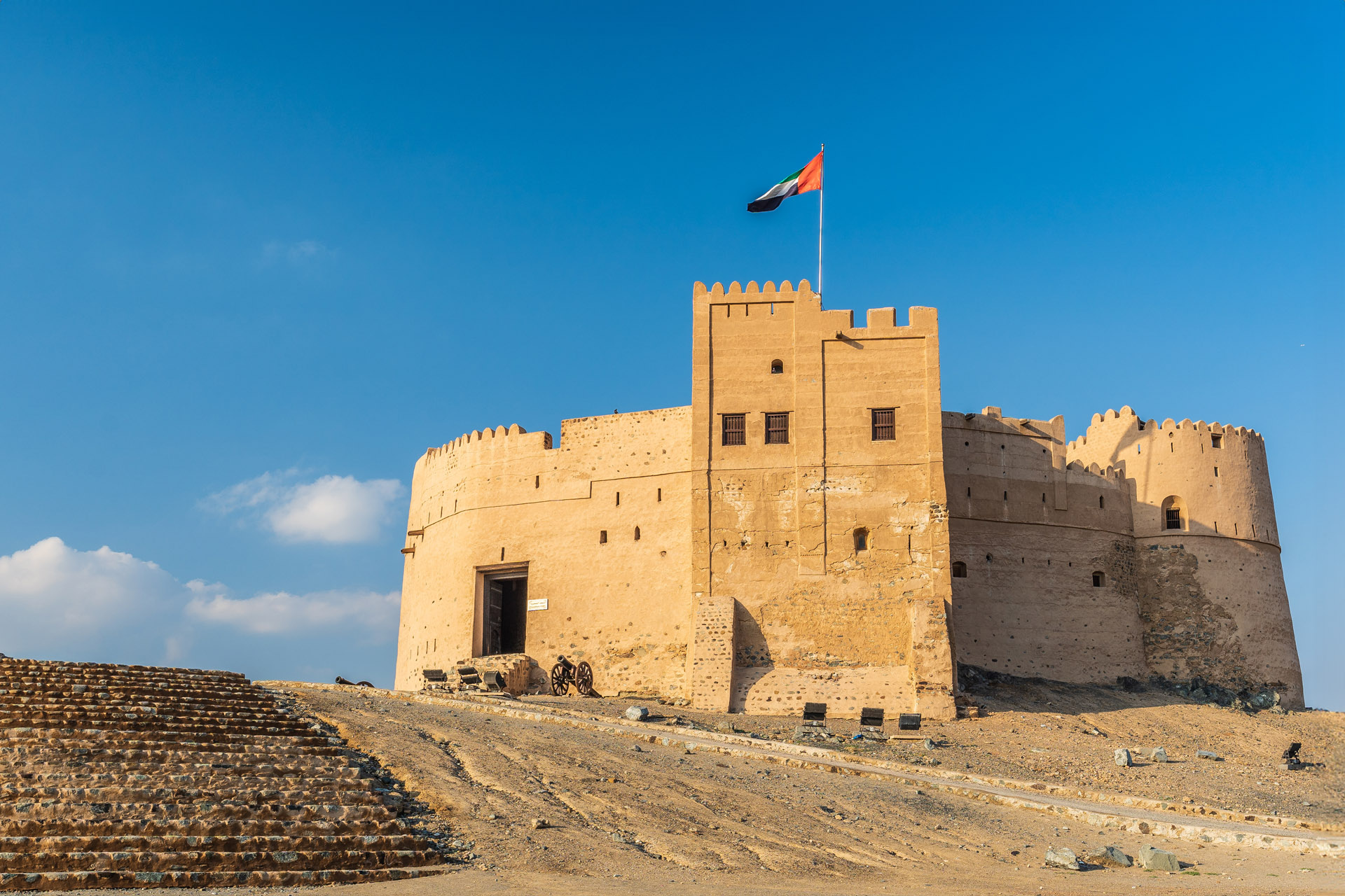 Photo looking up towards Fujairah Fort. it is a historic landmark in the UAE and is built from mud, bricks, and stones, featuring tall, sturdy walls with rounded and rectangular watchtowers. The UAE flag flies atop the central tower, and old cannons sit near the entrance. The fort, set against a clear blue sky, stands on a rocky elevation, showcasing its strategic and cultural significance.