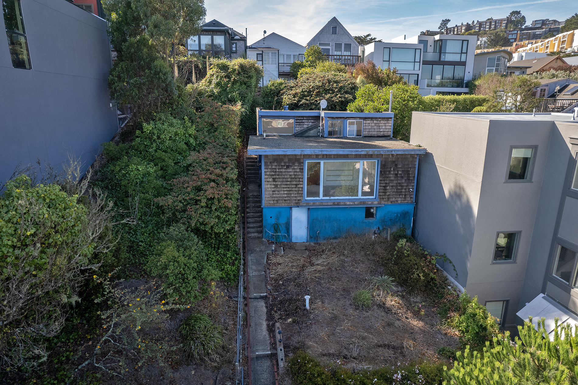 A small blue house sits at the top of narrow, inclined lot. There is greenery in the garden around it. 