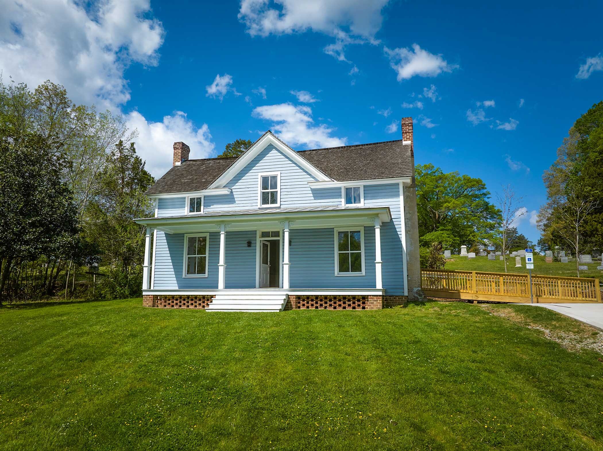 A bright blue two-story house with white trim sits on a grassy lawn. In the background, a small cemetery with headstones is visible to the right, behind a wooden fence and ramp.