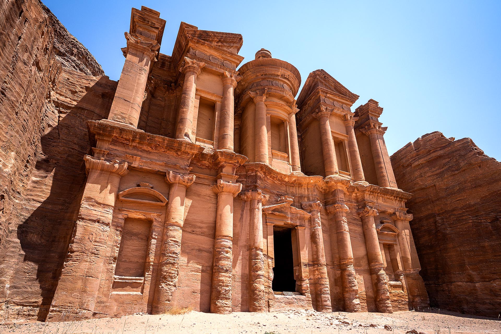 The image shows Ad-Deir, or the Monastery, in Petra, Jordan, a massive rock-cut monument carved from rose-colored sandstone. Its grand façade features columns, niches, and a central doorway topped by an urn-shaped crown. Sunlight highlights the warm tones of the stone against the deep blue sky, emphasizing the monument’s scale and harmony with the surrounding cliffs.